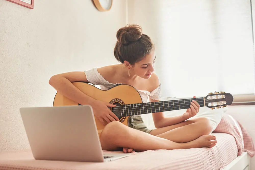 Girl learning acoustic guitar on bed with a laptop
