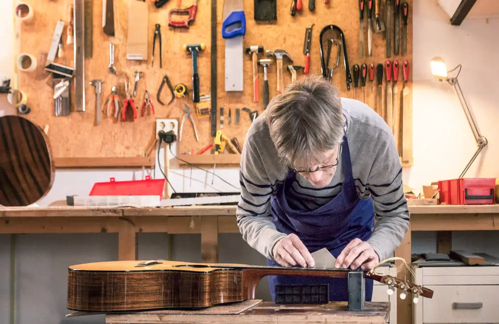 Luthier working on a workbench measuring and leveling the frets of a guitar with his tools in the background